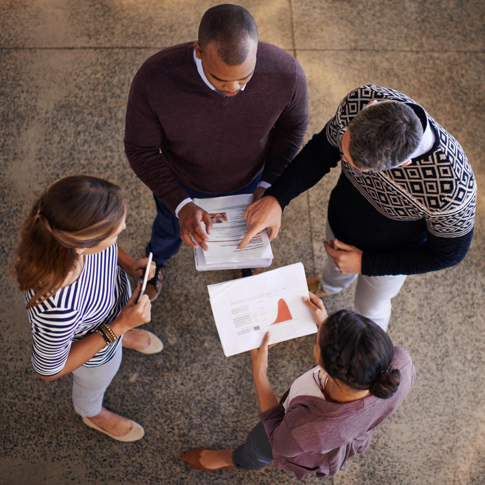 Four colleagues standing in a circle reviewing a printed report from overhead, one pointing to a page