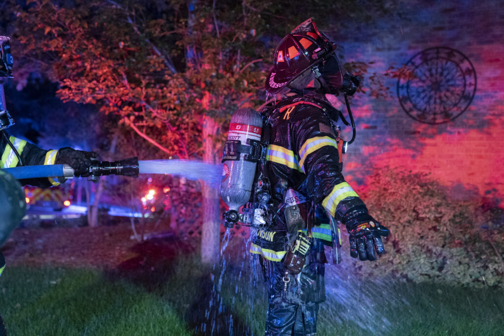 Firefighter being decontaminated with water hose on scene after a fire, turnout gear soaked and visibly contaminated, illustrating the importance of on-scene gear cleaning to reduce PFAS exposure.