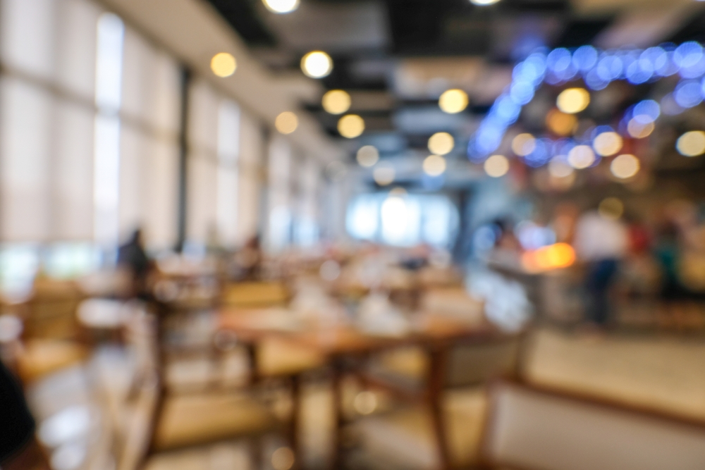 Blurred restaurant dining room with warm string lights and bokeh, empty tables and chairs in soft focus, staff visible near the counter in the background.